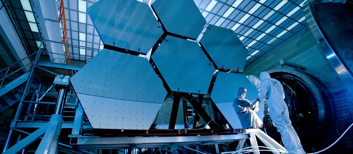 Engineers in protective suits work on telescopic mirrors in a high-tech lab.