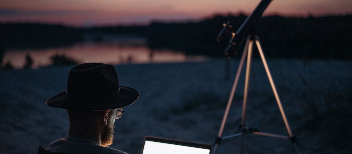 A man enjoying stargazing by a serene lake at twilight, using a telescope and laptop.