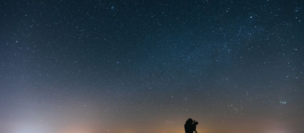 A person using a telescope under a vast, star-filled night sky with a silhouette effect.