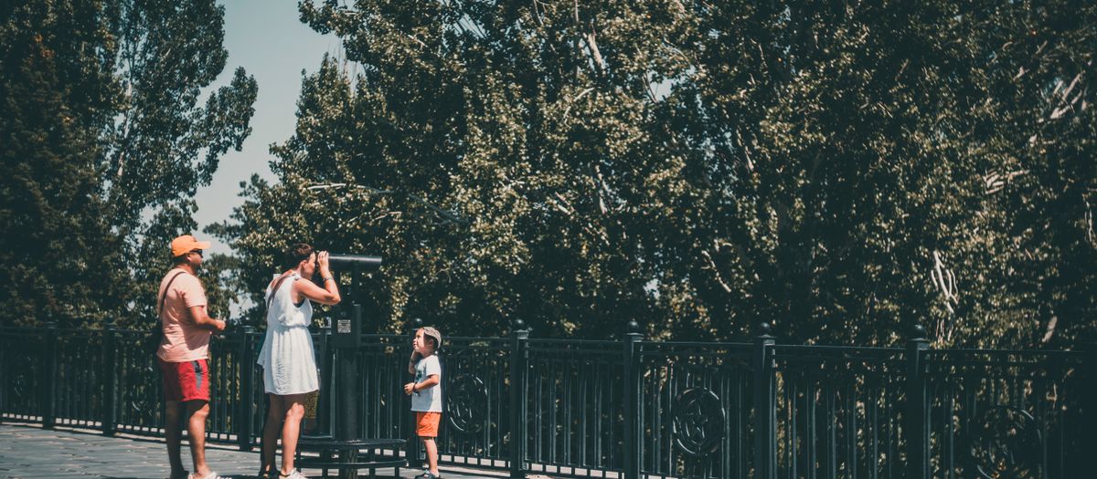 A family with a child uses a telescope in a park setting, surrounded by lush trees.