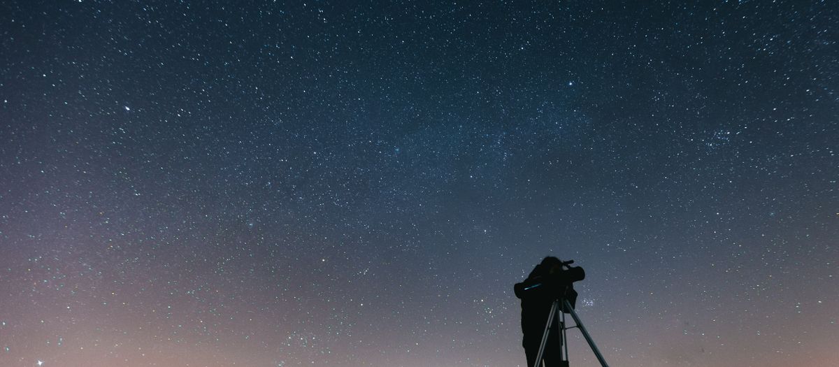 A silhouetted person using a telescope to observe the night sky filled with stars.